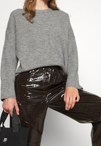 Femme portant un pull gris côtelé et un pantalon marron foncé brillant, tenant un sac fourre-tout noir avec des anses à motifs.