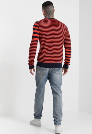 Mann mit tätowiertem Hals, der einen rot-navy gestreiften Pullover, hellblaue Jeans und weiße Turnschuhe trägt und mit dem Rücken zum Betrachter vor einem weißen Hintergrund steht.