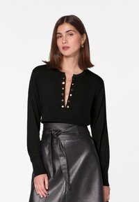 Zwart lange mouwen sweater met een diepe V-halslijn, gouden knopen en een decoratieve ketting. Dragen bij een hoogopgesneden zwarte leren rok.