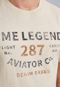 Béžové bavlněné tričko s kulatým výstřihem, na kterém jsou vytištěné černé, hnědé a šedé nápisy, včetně "Aviator Co." a "Let č. 287."