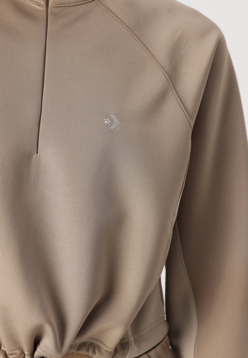 Top sportivo beige con un collo alto, tessuto texture e logo ricamato sul petto. Presenta un orlo arricciato e maniche raglan.