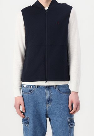 Personne portant une veste sans manches zippée bleu marine par-dessus une chemise blanche à manches longues et un jean cargo en denim bleu, debout devant un fond blanc.