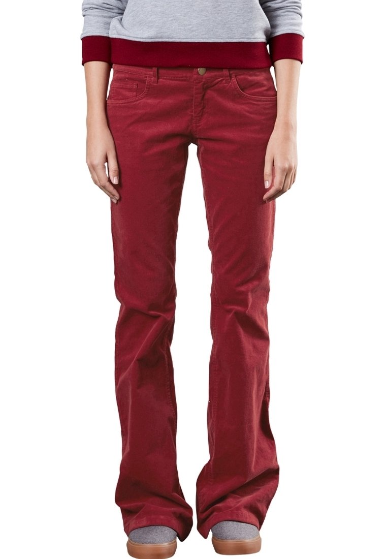 Personne portant un pantalon en velours côtelé rouge, des chaussures grises et un sweat-shirt gris avec des poignets rouges, debout sur un fond blanc.
