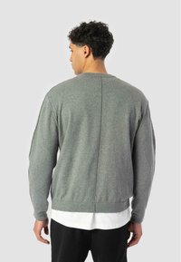 Hombre con cabello rizado que lleva una sudadera gris de manga larga sobre una camisa blanca, de espaldas contra un fondo claro y liso.