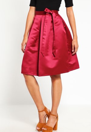 Femme portant une jupe en satin rouge longueur genou avec une ceinture nouée et des sandales marron à talons hauts, debout sur un fond blanc.