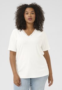 T-shirt blanc à manches courtes avec une texture côtelée, doté d'un décolleté en V orné de dentelle. Associé à un jean bleu clair.