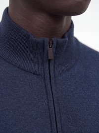 Gros plan d'une personne à la peau foncée portant un pull en maille côtelée bleu marine avec un col montant zippé.