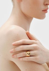 Anillo de color dorado con una textura ondulada, usado en un dedo que reposa sobre un hombro descubierto. Tonos de piel suaves destacados en un entorno simple.