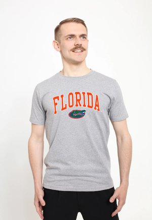 Homme avec moustache portant un t-shirt gris Florida et un pantalon noir, regardant légèrement vers le haut et sur le côté, souriant.