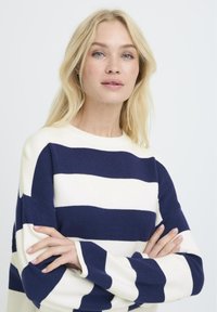 Suéter a rayas en colores azul marino y crema, con tela suave, cuello redondo y hombros caídos. Manga larga con puños doblados.