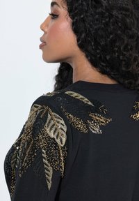 Top nero con intricate foglie ricamate in paillettes oro e nere sulle spalle, che mettono in risalto un design testurizzato.