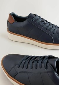 Sneakers in pelle blu navy con finitura testurizzata, caratterizzati da un accento sul tallone marrone e una suola in gomma bianca, distintivi per i lacci piatti e un design minimalista.