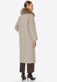 Langer beiger Mantel mit einem Kunstfellkragen, gerader Schnitt und strukturiertem Stoff. Das Modell trägt braune Hosen und High Heels.