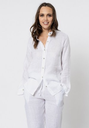 Vrouw met bruin haar, gekleed in een wit linnen overhemd met knopen en bijpassende witte linnen broek, staand met handen in de zakken.
