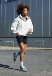 Giacca bianca con zip, pantaloni corti neri, scarpe da corsa grigie e calzini azzurri chiari; il modello sta correndo su un marciapiede con un edificio moderno grigio sullo sfondo.