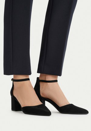 Pies calzados con tacones de bloque de ante negro con correa en el tobillo, combinados con pantalones negros recortados de corte sartorial, sobre un fondo neutro.