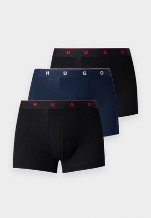 Svalnatý muž stojaci naboso, oblečený v čiernych boxerkách s červeným pásikom "HUGO", pozerá sa doprava na jednoduchom bielom pozadí.