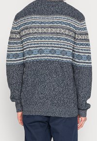 Homme portant un pull en tricot bleu marine à motifs et un pantalon bleu foncé, vu de dos sur un fond uni.