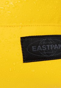 Tessuto impermeabile giallo con gocce d'acqua visibili. Toppa del logo Eastpak nera cucita sulla superficie. Texture e design durevoli.