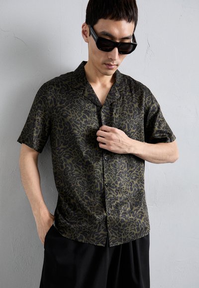 Camicia a maniche corte di colore verde scuro con un motivo astratto nero, caratterizzata da un colletto rilassato e chiusura frontale con bottoni, realizzata in un tessuto leggero e liscio.
