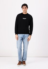 Schwarzer Hoodie mit weißem Text auf der Brust, kombiniert mit hellblauen Jeans und braunen Sneakers. Einfarbige Farben, lässige Passform.