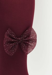 Vinröda strumpbyxor med en stor, texturerad rosett av genomskinligt material med små silverprickar, placerad på utsidan.
