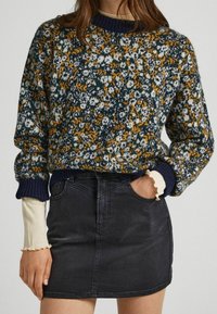 Gestrickter Pullover mit floralem Muster in Marineblau, Senfgelb und Weiß auf dunkelgrünem Hintergrund; kombiniert mit einem schwarzen Jeansrock.