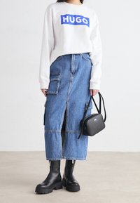 Felpa bianca con logo blu "HUGO", abbinata a una lunga gonna di jeans blu con tasche cargo e stivali neri fino al polpaccio. Borsa a tracolla nera.