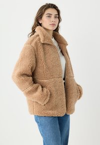 Beżowa, pluszowa kurtka z polaru z wysokim kołnierzem, zamkiem błyskawicznym i dużymi kieszeniami. Ozdobiona gładkim materiałem wzdłuż zamka i szwów.