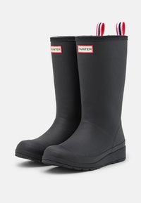 Bottes de pluie en caoutchouc noir avec une surface texturée, des tirants rayés rouge et blanc, et un marquage "HUNTER" bien en vue à l'avant.