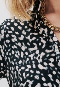 Camicia nera con un motivo astratto bianco e grigio, dotata di colletto e un collier dorato. Il tessuto appare liscio e leggero.