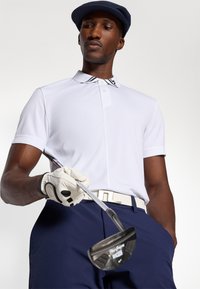 Hvit polo skjorte med en svart og hvit stripete krage, marineblå bukser, et hvitt belte og en hanske som holder en golfkølle. Blå caps fullfører antrekket.