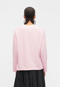 Άτομο με πλεξούδες που φοράει ριγέ πουκάμισο ροζ και άσπρο με μακριά μανίκια και σκουρόχρωμη πλισέ φούστα, γυρισμένο ανάποδα μπροστά σε ένα απλό φόντο.