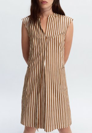 Donna che indossa un vestito senza maniche fino al ginocchio con righe verticali marroni e bianche e tasche frontali, in piedi davanti a uno sfondo semplice.