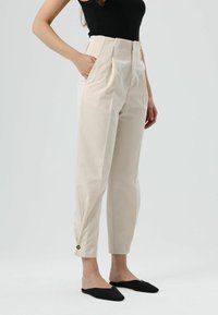 Beige hoch taillierte Hose mit vorderen Falten, Seitentaschen und einem Knopfdetail am Saum. Glatter Baumwollstoff mit maßgeschneiderter Passform.