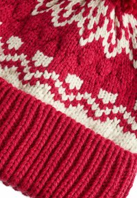 Bonnet tricoté rouge avec un motif géométrique blanc sur le dessus et une bande côtelée rouge en bas. Présente un pompon sur le dessus.