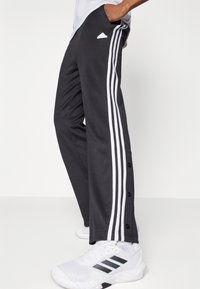 Svarta atletiska byxor med vita ränder längs sidorna, tryckknappar vid anklarna och en broderad logotyp på höften.