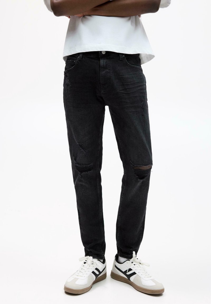 Personne portant un jean noir déchiré avec des baskets blanches et noires, debout, les bras croisés, devant un fond blanc uni.