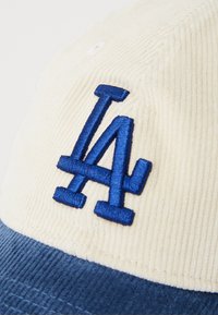 Kordfløyelslue med en kremfarget topp og en blå skjerm, dekorert med en blå brodert "LA" logo foran.