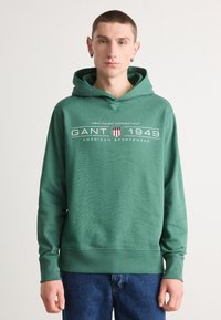 Felpa verde in cotone con polsini e orlo a costine, caratterizzata da una stampa grafica bianca e da un logo. Indossata sopra un paio di jeans in denim blu su uno sfondo semplice.