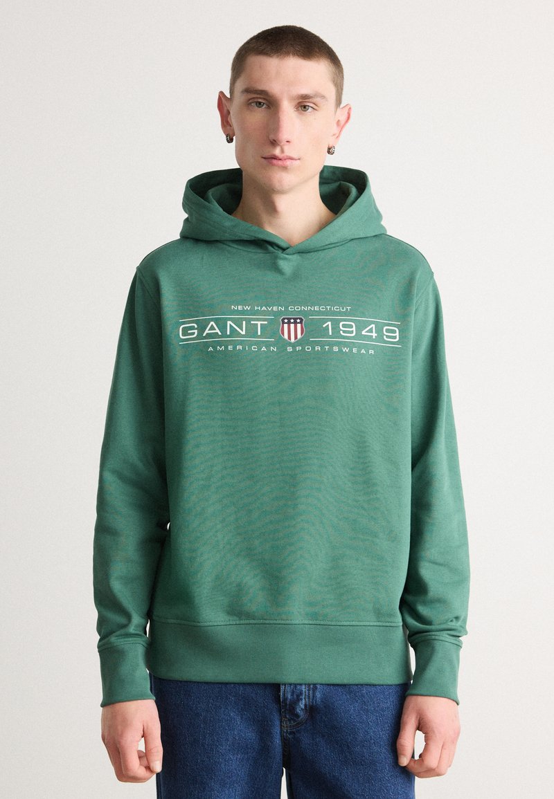 Felpa verde in cotone con polsini e orlo a costine, caratterizzata da una stampa grafica bianca e da un logo. Indossata sopra un paio di jeans in denim blu su uno sfondo semplice.
