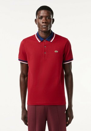 Camisa polo roja con cuello plano, con acentos en blanco y azul. Dos botones en el cuello y un logotipo verde en el lado izquierdo del pecho.