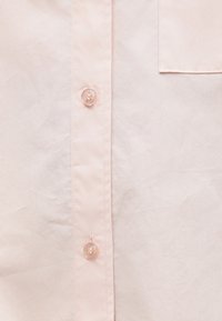 Devant de chemise rose clair affichant deux boutons transparents et une partie d'une poche poitrine avec des détails de couture visibles.