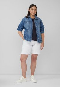 Frau steht mit Händen in den Taschen, trägt eine blaue Jeansjacke, ein schwarzes Oberteil, weiße Shorts und weiße Sneakers vor einem schlichten grauen Hintergrund.
