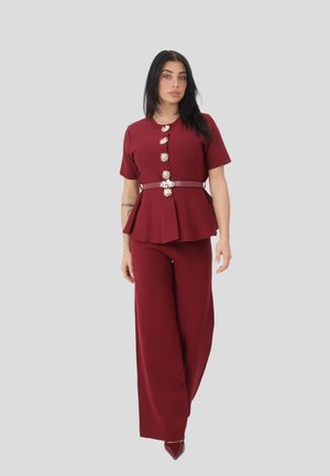 Donna che indossa un top peplum bordeaux a maniche corte con bottoni decorativi e pantaloni a gamba larga abbinati su uno sfondo grigio uniforme.
