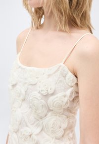 Γυναίκα που φοράει λευκό δαντελένιο φόρεμα με τρισδιάστατες λουλουδένιες εφαρμογές και λεπτούς λεπτούς ιμάντες, εμφανίζεται από τους ώμους μέχρι το μέσο του κορμού.