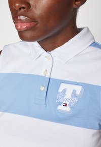 Camisa polo en rayas blancas y azules claras, con cuello, botonera y logo bordado de "Tommy" en blanco con un pequeño acento rojo y negro.