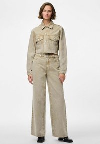 Veste en denim beige coupée avec deux poches poitrine, associée à un pantalon en denim beige à jambe large. Des chaussures noires complètent la tenue.