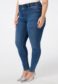 Jeggings en denim bleu avec une taille élastique, une jambe étroite et des détails de couture classiques. Associés à des escarpins argentés à bout pointu.