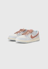 Nike sportbačiai su šviesiai pilku tinklo paviršiumi, oranžinėmis detalėmis, smėlio spalvos guma ir tekstūruota medžiaga. Šnervės atitinka oranžines detales.